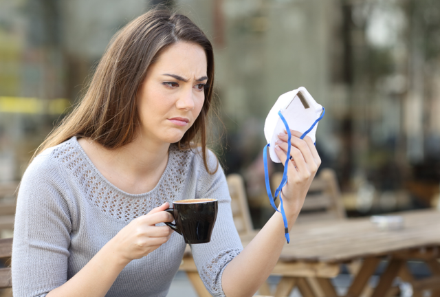 A woman sitting down having a coffee and staring at a facial mask in her other hand