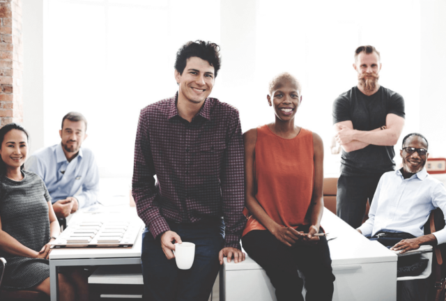 Employees in a boardroom all around a table. Two of them are standing in front of the table smiling