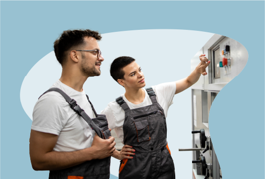 A younger employee is showing an older employee hot to do a task on a piece of industrial equipment. They are both matching wearing white t-shirts and brown coveralls